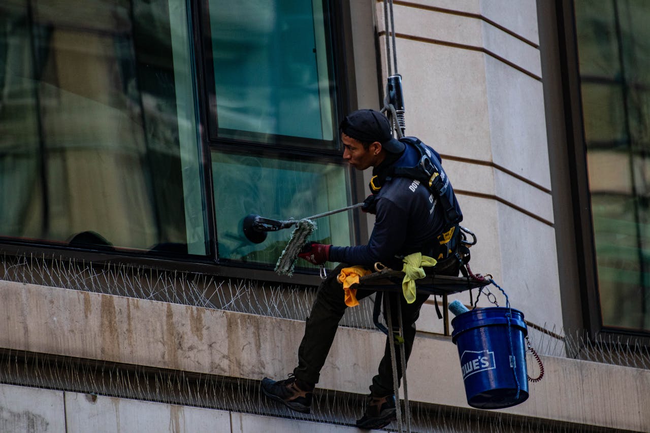 Man cleaning high-rise building windows using safety harness outdoors in daylight.